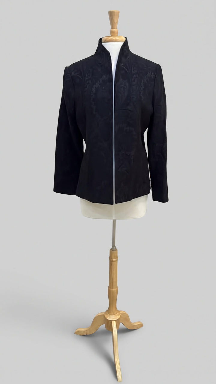 A black long-sleeve jacket with a subtle damask pattern, displayed on a cream-colored mannequin torso mounted on a wooden stand against a solid gray background.