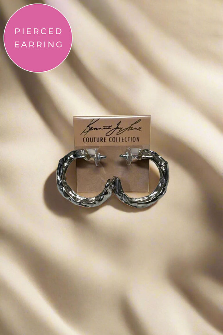 A pair of textured silver hoop earrings displayed on a small branded card against draped beige fabric. A pink circular badge in the top left reads 'PIERCED EARRING.