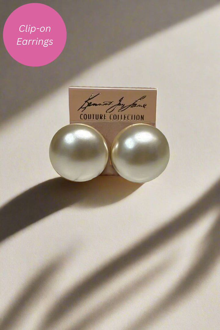 A pair of round, pearl-like clip-on earrings in a warm off-white/ivory shade, displayed on a small beige card labeled 'COUTURE COLLECTION'. The image is styled with dramatic, directional shadows on a light beige background, and a pink circular graphic overlay in the top-left corner reading 'Clip-on Earrings'.