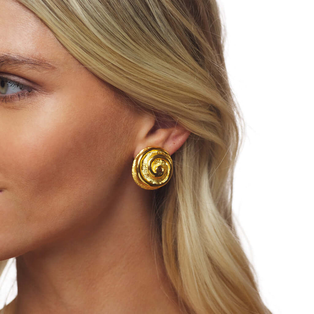 A close-up of a woman's ear wearing a textured, spiral-shaped gold clip-on earring against a plain white background.