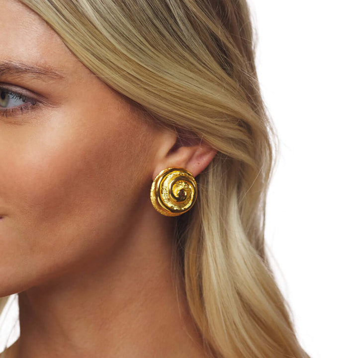 A close-up of a woman's ear wearing a textured, spiral-shaped gold clip-on earring against a plain white background.