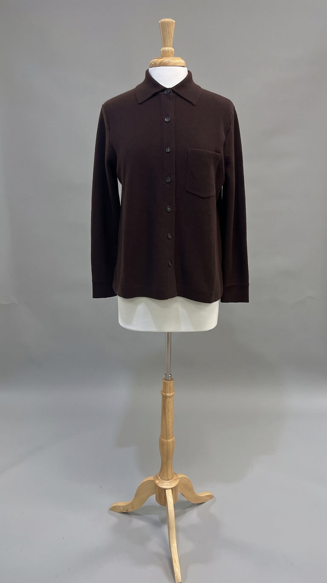 A dark brown long-sleeve button-up shirt with a chest pocket, displayed on a white mannequin torso mounted on a wooden stand against a gray studio background.