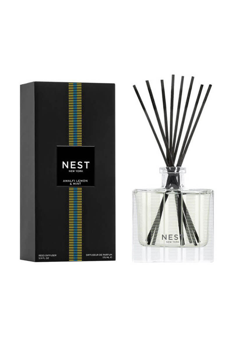 A reed diffuser bottle with black reeds next to its black packaging box, both featuring the NEST New York logo and a vertical blue and yellow stripe design, set against a plain white background.