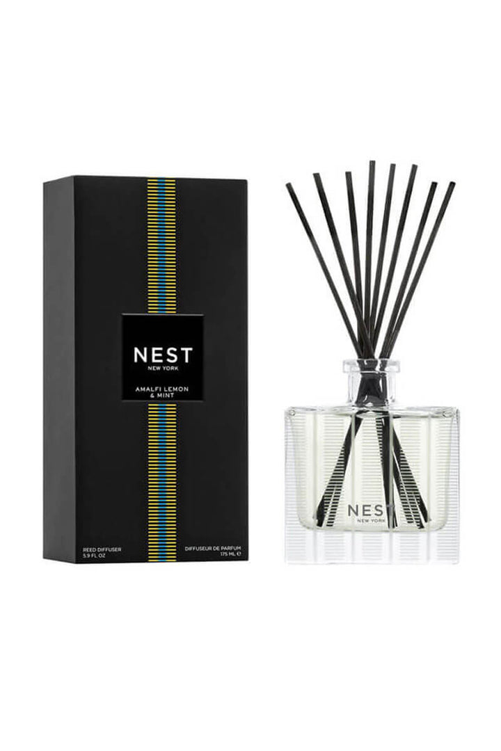 A reed diffuser bottle with black reeds next to its black packaging box, both featuring the NEST New York logo and a vertical blue and yellow stripe design, set against a plain white background.