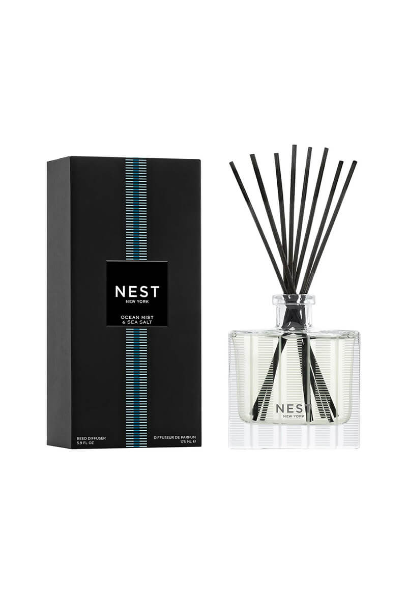 A reed diffuser bottle with black reeds next to its black packaging box, both featuring the NEST New York logo and a blue vertical stripe design, on a clean white background.