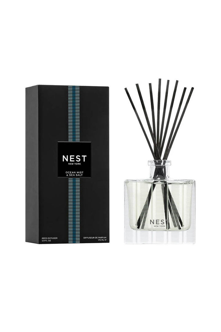 A reed diffuser bottle with black reeds next to its black packaging box, both featuring the NEST New York logo and a blue vertical stripe design, on a clean white background.