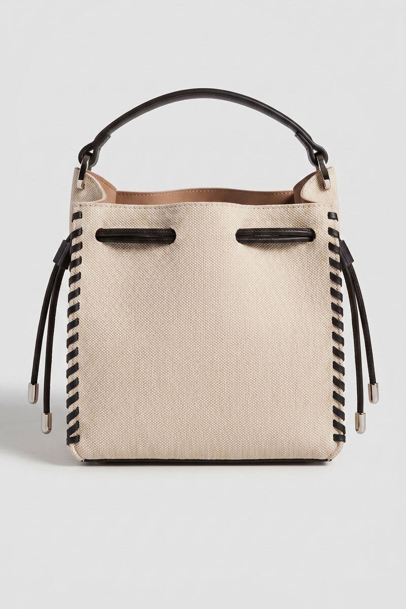 A structured canvas bucket bag in beige with black trim, a black top handle, and black braided strap details hanging from the sides, shown against a light gray background.