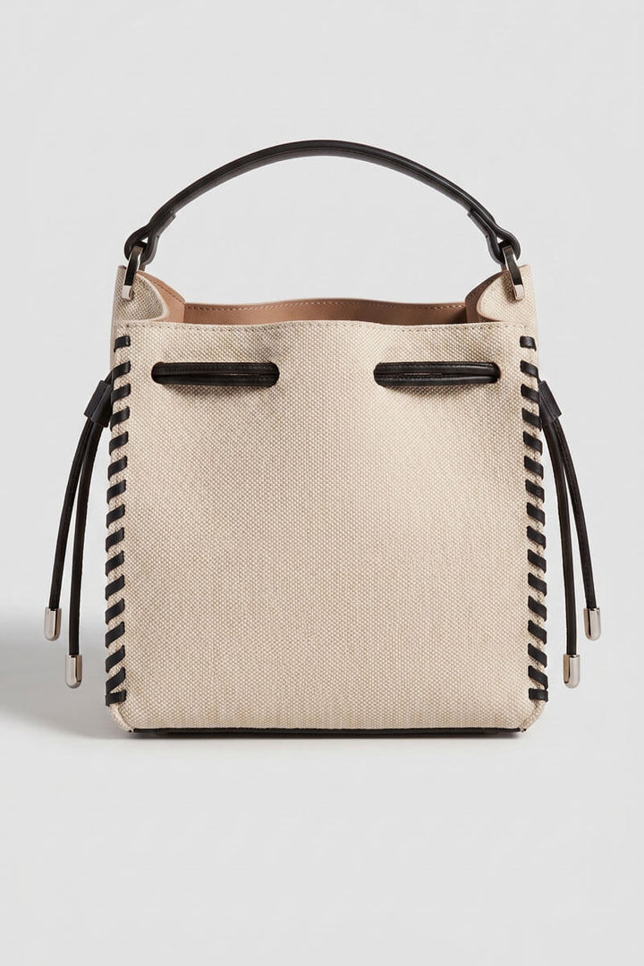 A structured canvas bucket bag in beige with black trim, a black top handle, and black braided strap details hanging from the sides, shown against a light gray background.