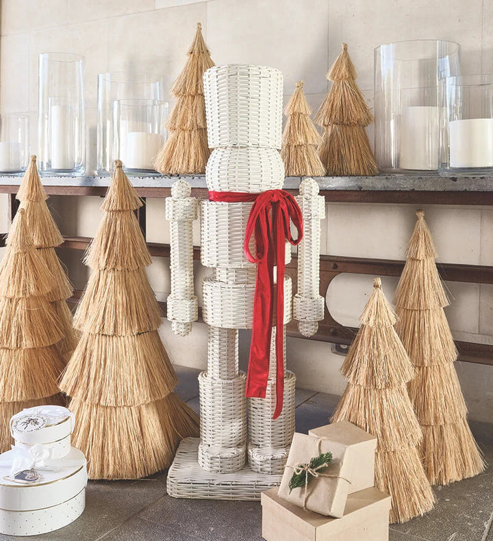 A white woven nutcracker holiday decoration with a bright red ribbon bow, standing in front of a shelf with straw Christmas trees and glass candle holders.