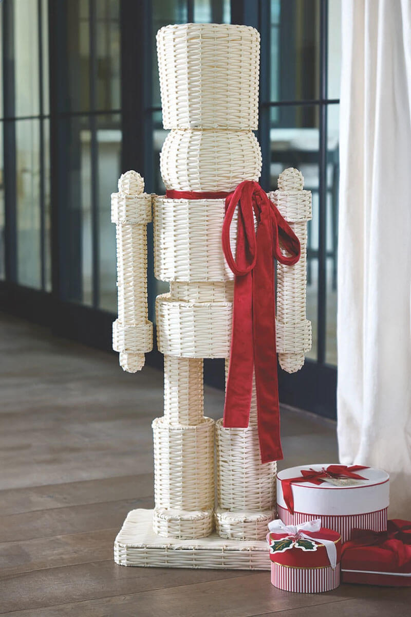 A tall, cream-colored woven rattan nutcracker decoration with a bright red ribbon bow, standing on a tiled floor next to wrapped gift boxes.